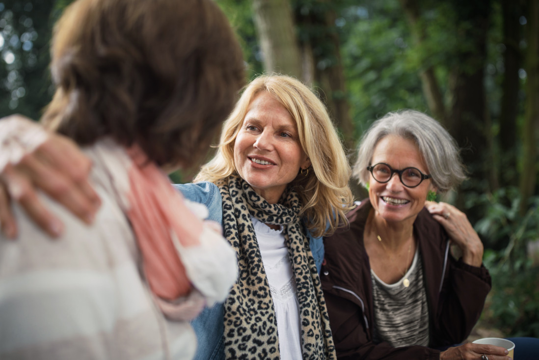 Drei Frauen, etwa Anfang/Mitte 50, stehen eng beieinander. Die Frau im Vordergrund sieht man von hinten. Die beiden anderen Frauen schauen sie lächelnd an. Die Frau in der Mitte hat die Arme um die beiden anderen gelegt. Auf ihr liegt der Bildfokus. Im Hintergrund sind grüne Bäume zu sehen.