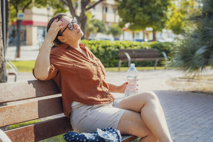Eine Frau sitzt bei heißen Temperaturen auf einer Bank im Freien. Sie hält in der linken Hand eine Flasche Wasser und hat die Augen geschlossen.