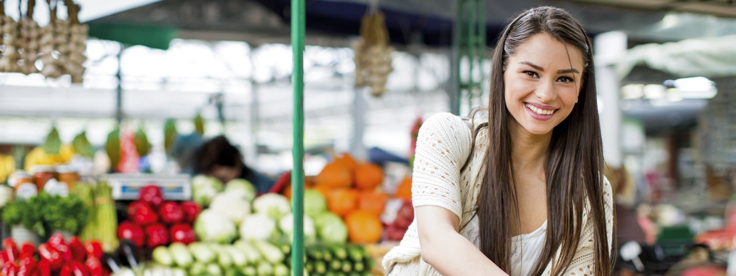 Eine junge brünette Frau kauft auf einem Markt Gemüse ein und lächelt.