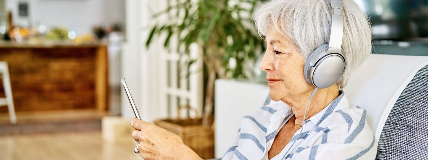 Eine Seniorin hört über ihre Kopfhörer den AOK Podcast Pflege. Dabei schaut sie auf ihr Smartphone und lächelt.