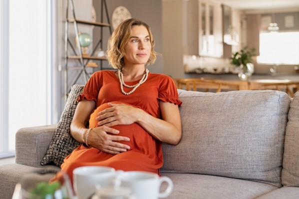 Eine etwa 40-jährige schwangere Frau sitzt nachdenklich auf einem Sofa.