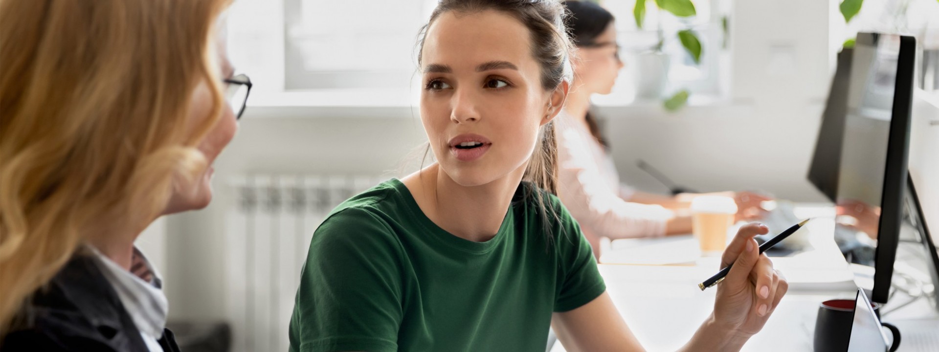 Eine etwa 30 jährige Frau unterhält sich an ihrem Schreibtisch im Büro mit ihrer Kollegin. Sie haben einen Laptop vor sich stehen.