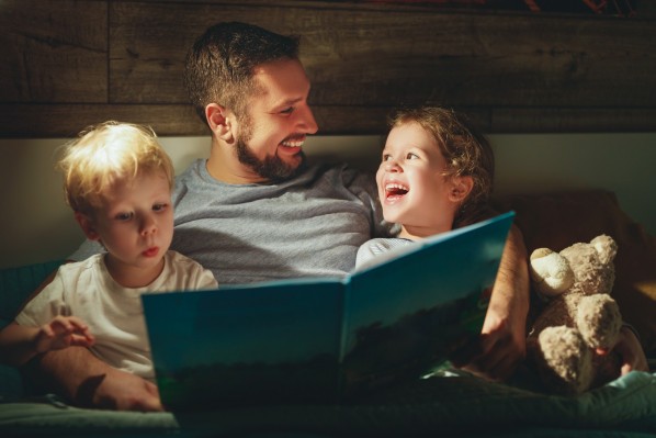 Vater lacht beim Vorlesen eines Buches mit Tochter und Sohn am Abend. Der Sohn schaut erstaunt auf den Inhalt des Buches.