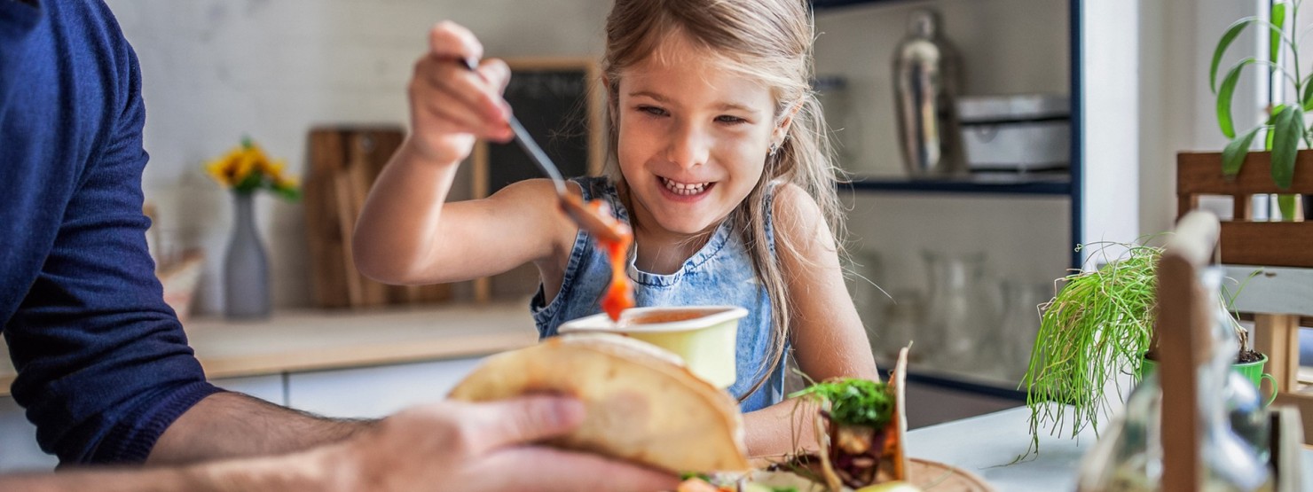 Ein circa 7-jähriges Mädchen macht mit ihrem Vater frische Tacos. Sie nimmt mit einem Löffel eine rote Soße aus einer Schüssel. Ihr Vater hält ihr ein Taco zum Befüllen bereit.