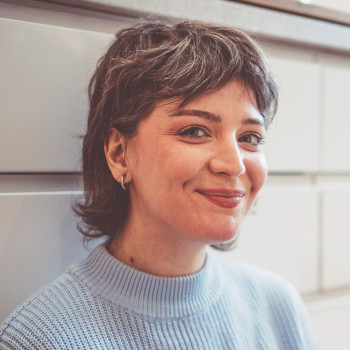 Nahaufnahme einer etwa 30-jährigen Frau mit Kurzhaarfrisur. Sie sitzt angelehnt an Unterschränken einer Küche und lächelt mit geschlossenem Mund.