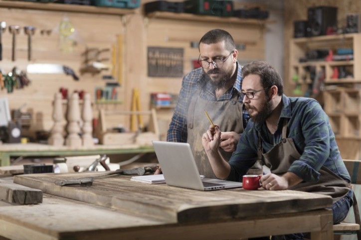 Zwei Handwerker befinden sich in einer Werkstatt und schauen interessiert auf einen Laptop.