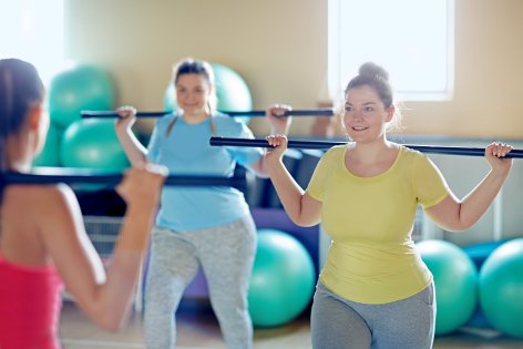 Drei Frauen machen gemeinsam Rehasport mit einer Stange auf den Schultern.