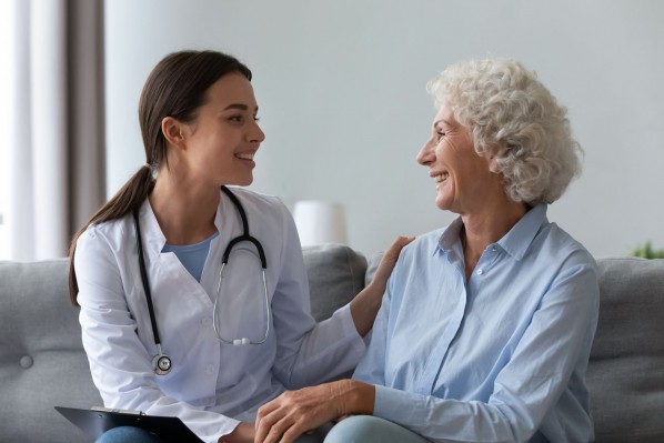 Eine Seniorin sitzt im Behandlungszimmer auf einem Sofa. Neben ihr sitzt eine junge Ärztin. Beide lächeln sich gegenseitig an. Die Ärztin hat ihr linke Hand auf der Schulter der älteren Frau gelegt.