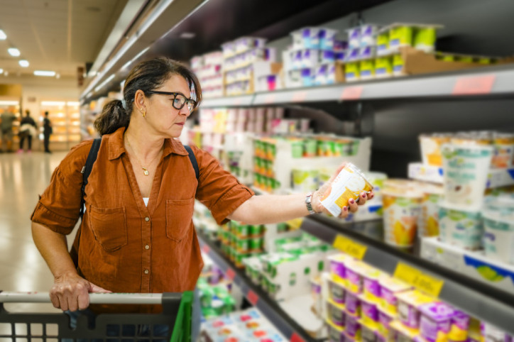 Eine etwa 50-jährige Frau läuft mit einem Einkaufswagen durch den Supermarkt und prüft die Rückseite von Produkten.