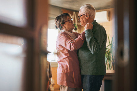 Ein Paar, beide Anfang bis Mitte 70, tänzeln gemeinsam sanft in einem Zimmer. Sie blicken einander liebevoll an. Man sieht sie durch einen Spalt in einer leicht geöffneten Holztür mit Glasscheiben.