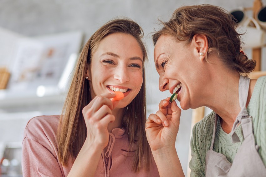 Eine circa 50-jährige Mutter und ihre circa 20-jährige Tochter naschen je eine Scheibe Gemüse, während sie gemeinsam kochen.