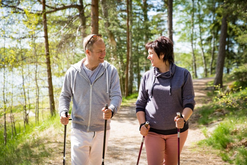 Ein etwa 50 jähriges Pärchen geht durch einen Wald mit Stöckern laufen. Beide schauen sich gegenseitig an und lächeln.