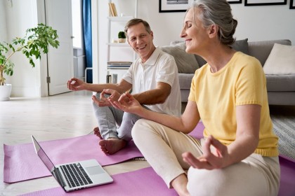 Ein älteres Pärchen sitzt auf zwei rosa Yogamatten im Wohnzimmer. Vor ihnen steht ein laptop. Beide machen Yoga und lächeln sich an.