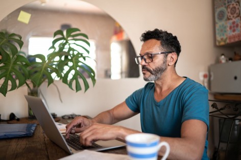 Ein etwa 40 jähriger Mann sitzt an einem Tisch und tippt auf seinem Laptop.