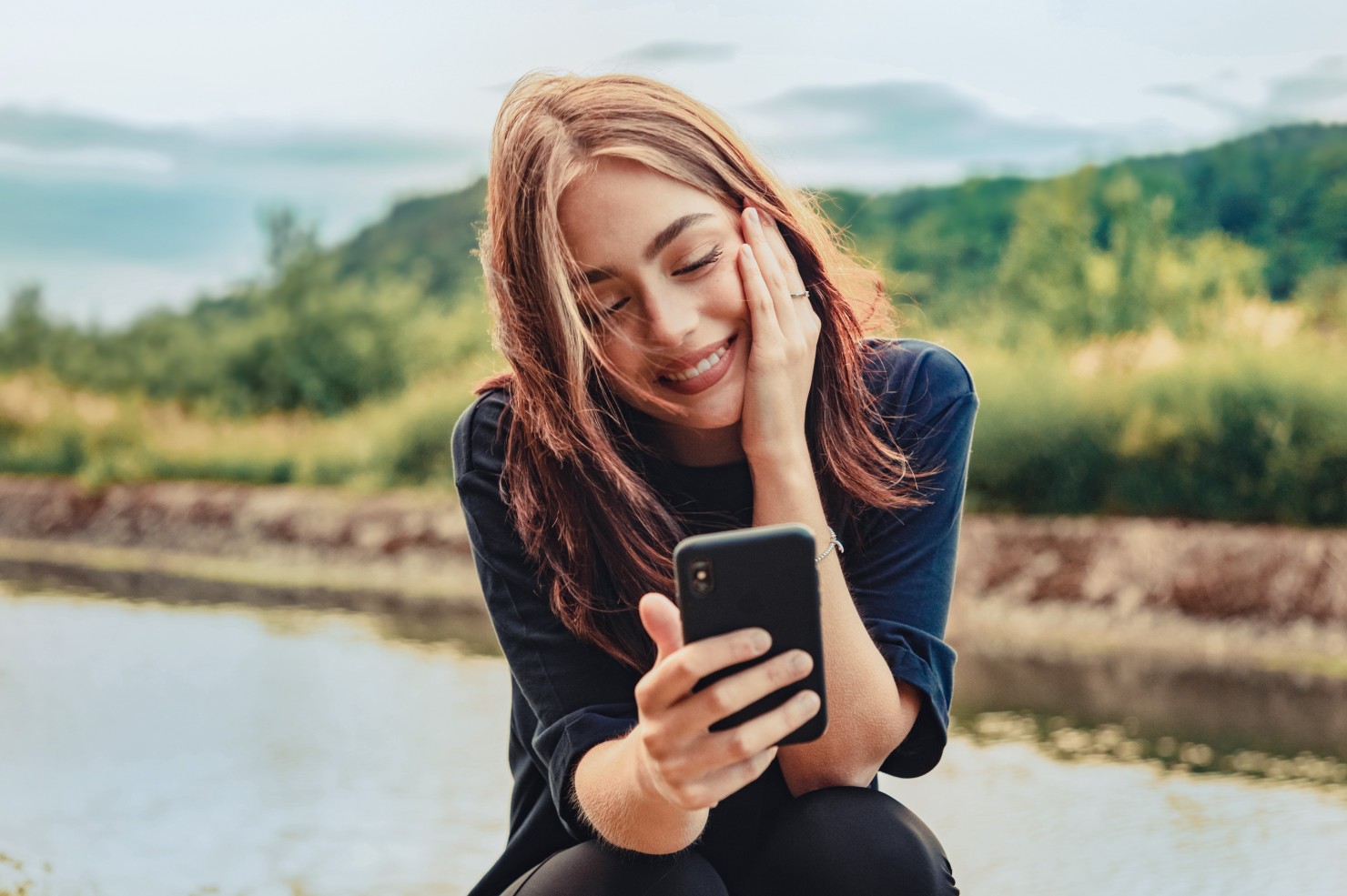 Eine etwa 30-jährige Frau sitzt an einem Fluss und schaut auf ihr Handy.