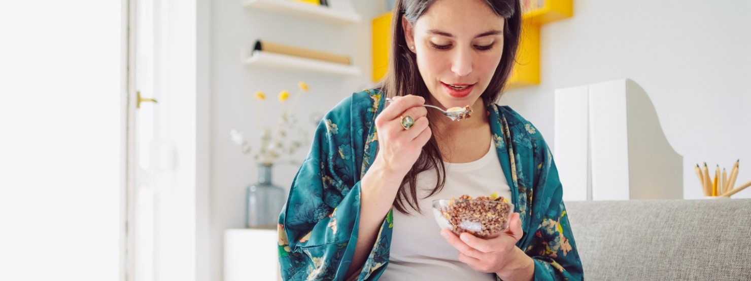 Eine etwa 30-jährige Frau sitzt auf dem Sofa und isst Müsli.