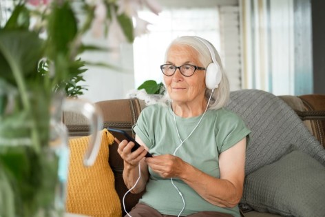 Eine Seniorin sitzt auf einem Sofa und hört über Kopfhörer einen Podcast.