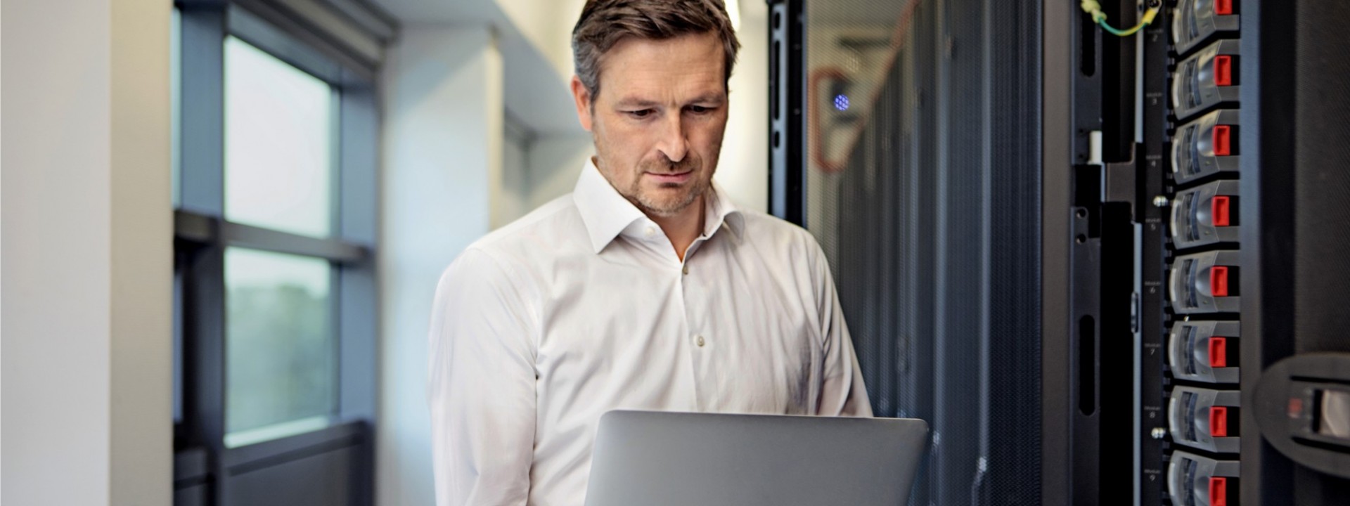 Ein etwa 40 jähriger Mann in weißem Hemd steht mit einem Laptop in der Hand vor einem Server.
