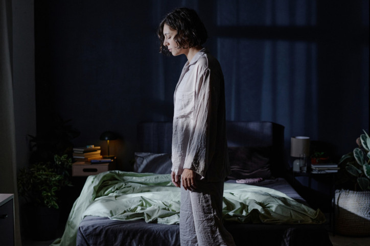 Eine Frau steht schlafwandeln in ihrem Schlafzimmer vor dem Bett. Dabei hat sie die Augen geschlossen.