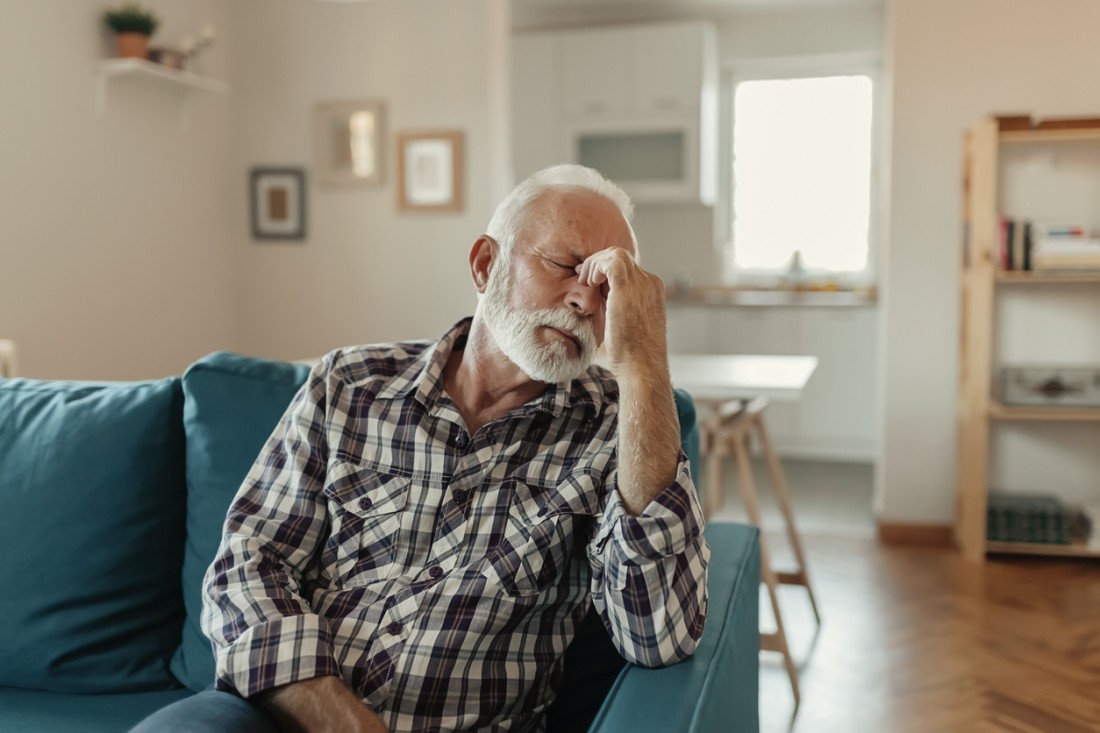 Senior sitzt auf dem Sofa und hält sich den Kopf.