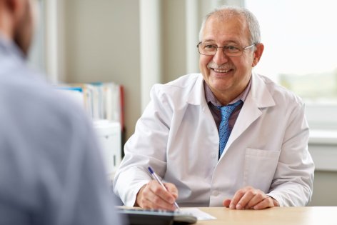 Ein etwa 60 jährige Arzt sitzt an seinem Beratungstisch und berät einen Patienten. Dabei lächelt er.