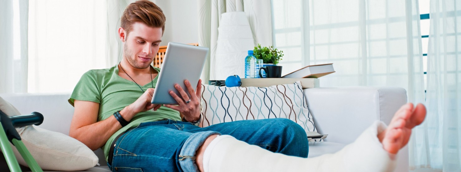 Ein junger Mann sitzt mit gebrochenem Bein auf dem Sofa und hat ein Tablet in der Hand.