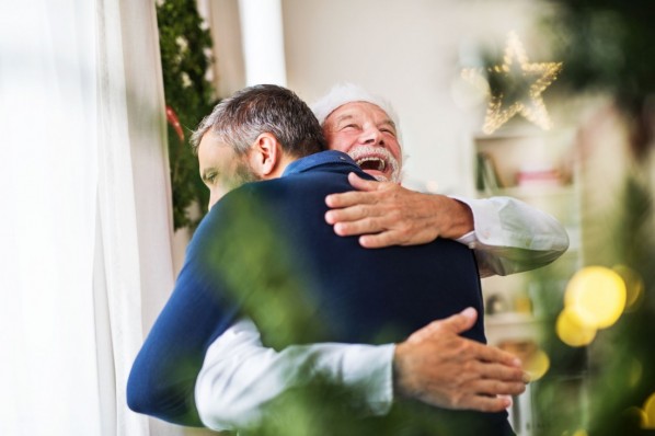 Ein etwa 70-jähriger Mann umarmt am Weihnachtsfest seinen etwa 45-jährigen Sohn. Sie stehen neben dem erleuchteten Weihnachtsbaum. 