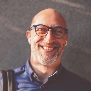 Nahaufnahme eines Mannes, etwa 45 Jahre alt, mit kahlem Kopf und kurzem, angegrautem Bart. Er trägt eine dunkelblaue Brille, ein dunkelblaues Hemd und weiße Bluetooth-Kopfhörer ihn den Ohren. Er lacht amüsiert mit offenem Mund und sichtbaren Zähnen. Lachfalten bilden sich um seine Augen.