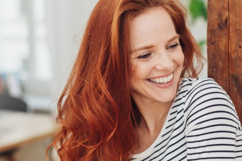 Eine rothaarige junge Frau hat einen gestreiften Pullover an, sitzt in einer Wohnung und lächelt.