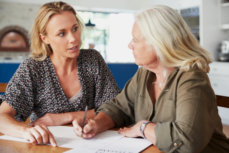 Zwei Frauen sitzen in einer Wohnung am Tisch und unterhalten sich. Die rechte Frau hält in der rechten Hand einen Stift und macht Notizen.