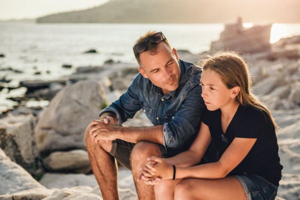 Ein Vater unterhält sich mit seiner etwa 12 jährigen Tochter. Dabei sitzen sie am steinigen Strand einer Bucht.