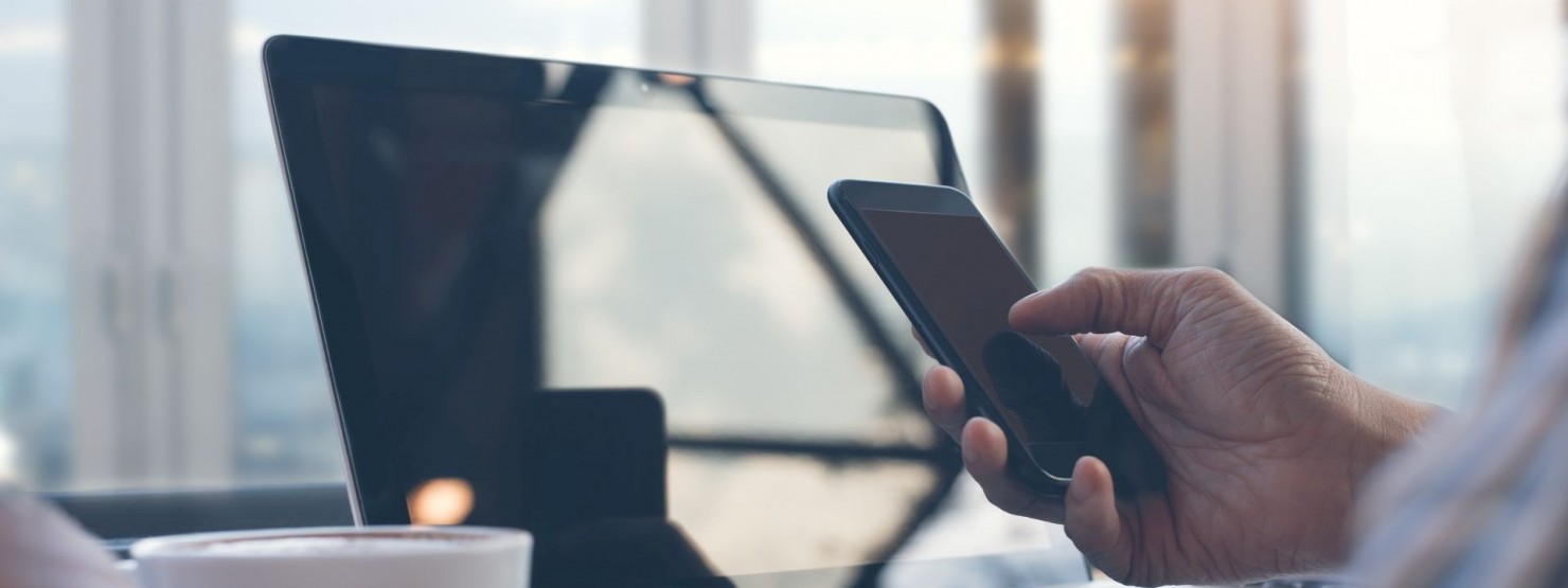 Eine Person hält in der rechten Hand ein Handy und sitzt vor einem Laptop.