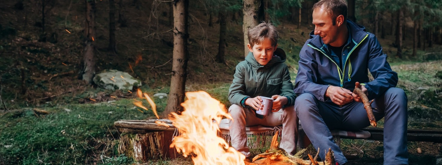 Ein Vater sitzt mit seinem etwa 10 jährigen Sohn an einem Lagerfeuer im Wald.