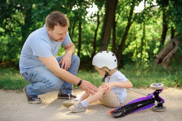 Ein Vater prüft das Bein seines etwa 3-jährigen Sohnes, der mit seinem Roller hingefallen ist.