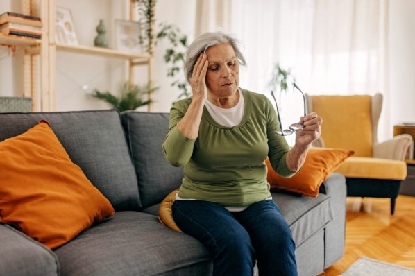 Eine Seniorin hält sich den Kopf während sie auf einem Sofa sitzt.