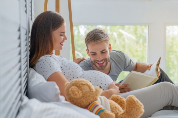 Eine etwa 30-jährige Schwangere liegt auf dem Bett und streichelt ihren Babybauch. Ihr Partner hat ein Buch in der Hand und liest dem Ungeborenen etwas vor.