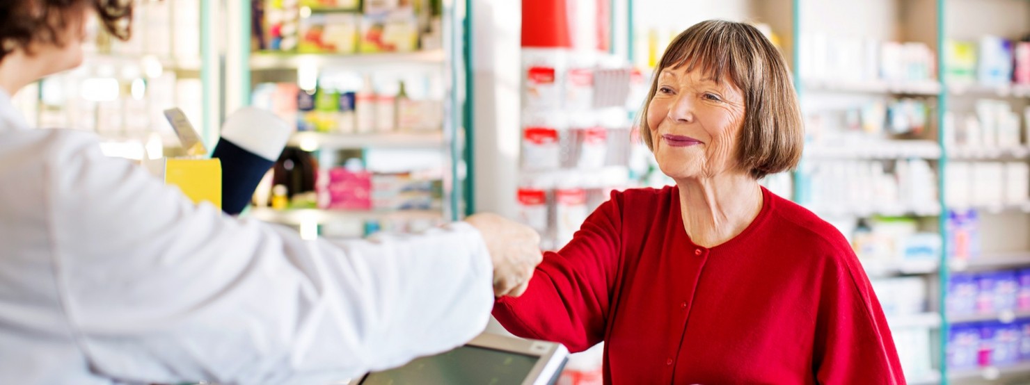 Eine Seniorin in roten Pullover steht in einer Apotheke und lässt sich von einer Apothekerin beraten. Sie lächelt.