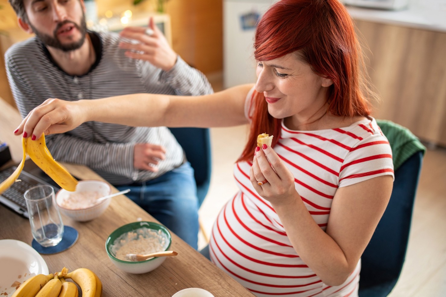 Eine etwa 30-jährige Schwangere und ihr Mann sitzen am Frühstückstisch. Sie isst eine Banane.