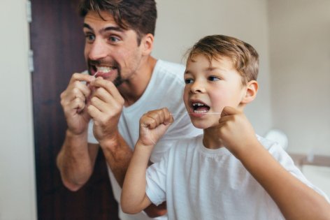Ein etwa 6 jähriger Junge steht mit seinem Vater im Badezimmer. Beide pflegen mit Zahnseide ihre Zähne.