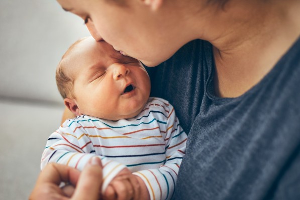 Eine junge Mutter hält ihr wenige Wochen altes Baby im Arm und küsst dessen Stirn.