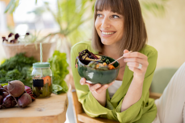 Young cheerful woman eats vegetarian lunch in bowl, sitting by the table full of fresh food ingredients indoors. Healthy lifestyle and wellness concept