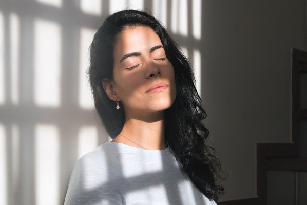 Eine etwa 30 jährige Frau lehnt mit geschlossenen Augen an einer Wand. Sie genießt die Sonnenstrahlen, die durch das Fenster scheinen.