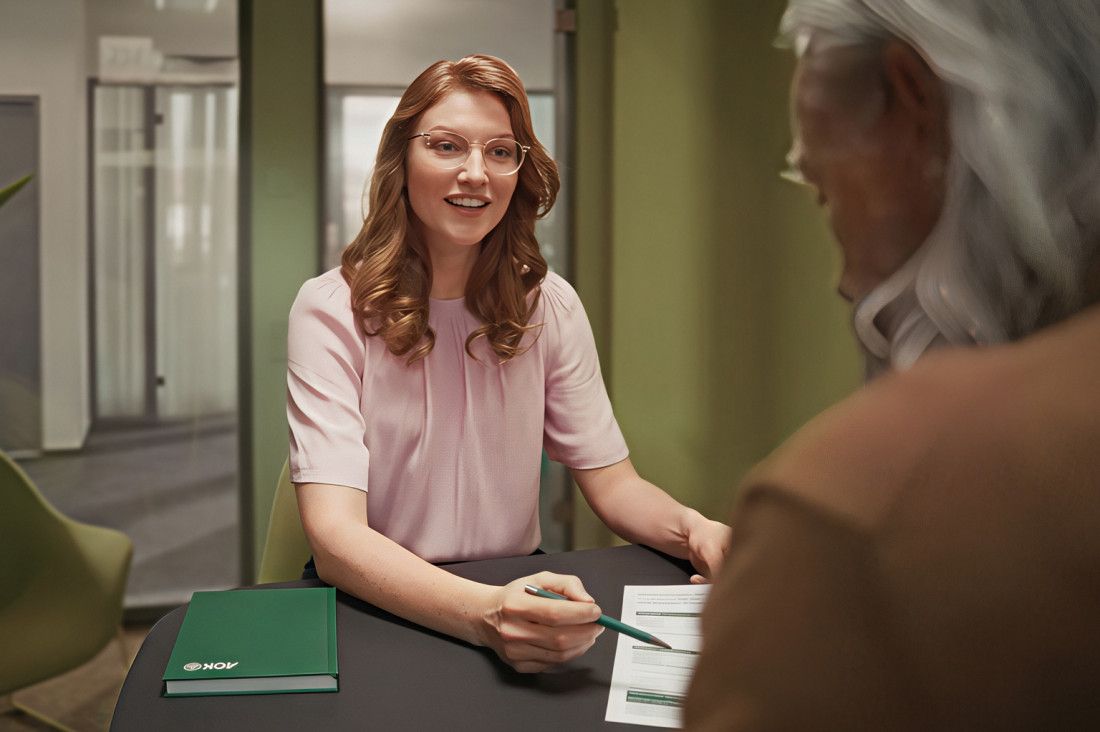 Eine etwa 30-jähriger Pflegeberaterin der AOK Sachsen-Anhalt sitzt einem etwa 75-jährigen Mann in einem Beratungsgespräch gegenüber. Man sieht sie offen lächelnd etwas erklären, dabei zeigt Sie mit deinem Bleistift auf ein Dokument. Sie befinden sich in einem Beratungsraum eines Kundencenters.