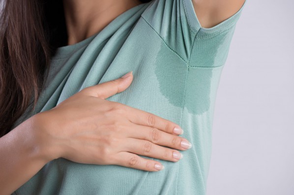 Eine Frau hebt ihren linken Arm. An ihrem T-Shirt zeichnet sich unter der Achsel ein Schweißfleck ab. 