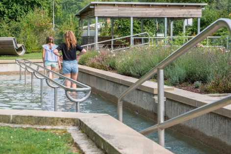 Junge Frau läuft barfuß durch einen schmalen, flachen Wasserlauf in einem Kiesbett.
