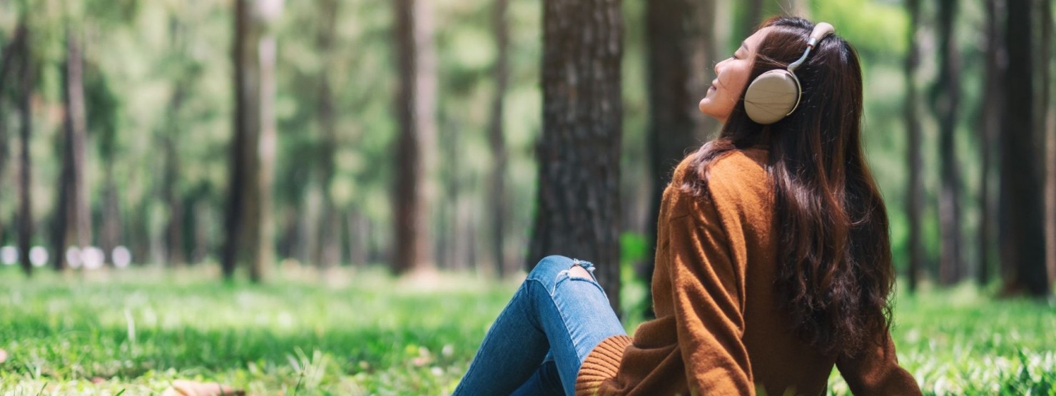 Eine etwa 25 jährige Frau sitzt mit Kopfhörern im Wald und genießt die Sonnenstrahlen.