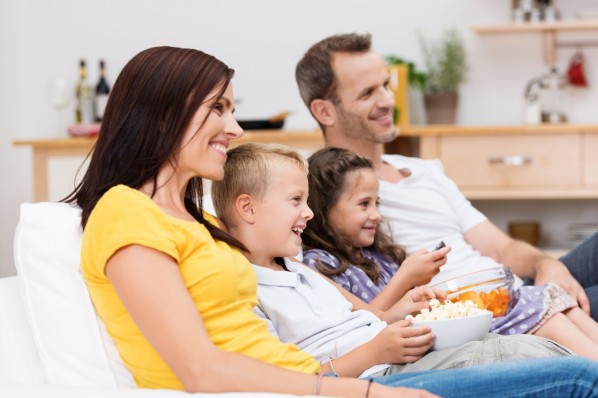 Eine vierköpfige Familie sitzt gemeinsam auf einer Couch und schaut lächelnd auf den Fernseher. Die Kinder haben Snacks auf ihrem Schoß.