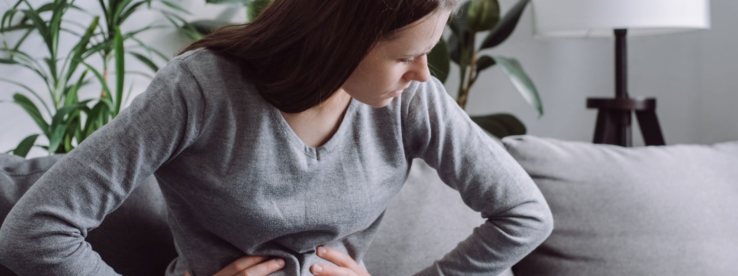 Junge Frau Mitte 30 sitzt auf der Couch. Sie hält ihren schmerzenden Bauch. 