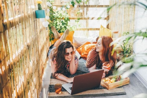 Zwei junge Frauen liegen auf einem Balkon in der Sonne, lachen und schauen auf einen Laptop.