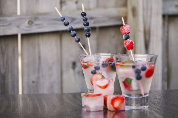 Auf einem Gartentisch stehen zwei gefüllte Wassergläser. Darin enthalten sind fruchtige Eiswürfel. Außerdem befinden sich in den Getränken frische Heidelbeeren und Erdbeeren.